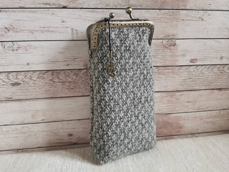 Puede incluir: Un estuche rectangular hecho de tela de tweed gris y blanca con un patr&oacute;n de diamantes texturizado. El estuche cuenta con un cierre de marco de metal color lat&oacute;n antiguo y un peque&ntilde;o dije colgante con forma de luna creciente y estrella.