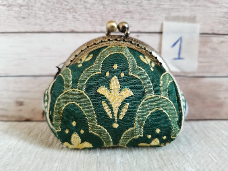 May include: A small coin purse featuring a dark green fabric with a repeating yellow floral pattern. The purse has a metallic bronze-toned kiss-lock clasp closure at the top. A small white square tag with the handwritten number 1 is visible.