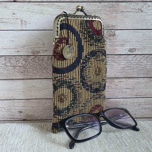May include: An eyeglasses case with a decorative pattern in shades of gold, navy, and burgundy, with a metal clasp. Black-framed eyeglasses rest in front of the case. The case has a small metal charm attached.