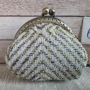 May include: A coin purse featuring a white woven fabric with a chevron pattern in metallic gold, green, and purple threads. The purse has an antique brass-toned metal frame with a double ball clasp closure and a small tag labelled with the number 6.