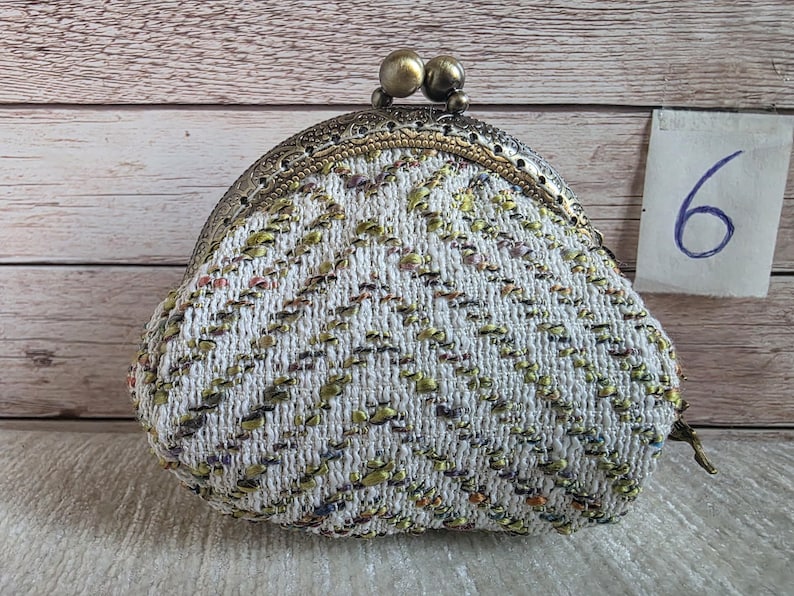May include: A coin purse featuring a white woven fabric with a chevron pattern in metallic gold, green, and purple threads. The purse has an antique brass-toned metal frame with a double ball clasp closure and a small tag labelled with the number 6.