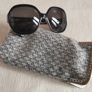 Puede incluir: Gafas de sol negras de gran tama&ntilde;o con lentes oscuras sobre un estuche rectangular para gafas de tela de tweed gris y blanco. El estuche cuenta con un cierre met&aacute;lico de color bronce con un marco texturizado y un exterior de tejido suave.