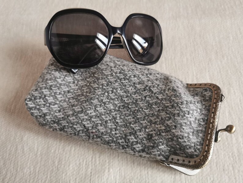 Puede incluir: Gafas de sol negras de gran tama&ntilde;o con lentes oscuras sobre un estuche rectangular para gafas de tela de tweed gris y blanco. El estuche cuenta con un cierre met&aacute;lico de color bronce con un marco texturizado y un exterior de tejido suave.