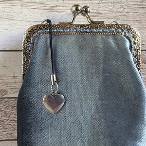 Puede incluir: Un bolso de terciopelo gris plateado con un marco de metal ornamentado y grabado que cuenta con un cierre de boquilla. Una correa de cord&oacute;n negro est&aacute; sujeta al marco, de la cual cuelga un peque&ntilde;o dije liso en forma de coraz&oacute;n plateado.