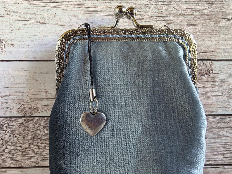 Puede incluir: Un bolso de terciopelo gris plateado con un marco de metal ornamentado y grabado que cuenta con un cierre de boquilla. Una correa de cord&oacute;n negro est&aacute; sujeta al marco, de la cual cuelga un peque&ntilde;o dije liso en forma de coraz&oacute;n plateado.