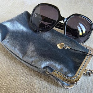 Puede incluir: Un par de gafas de sol negras de gran tama&ntilde;o sobre un estuche rectangular de color azul met&aacute;lico. El estuche cuenta con un cierre de boquilla plateado con un patr&oacute;n grabado ornamentado y un peque&ntilde;o dije de coraz&oacute;n plateado colgante.