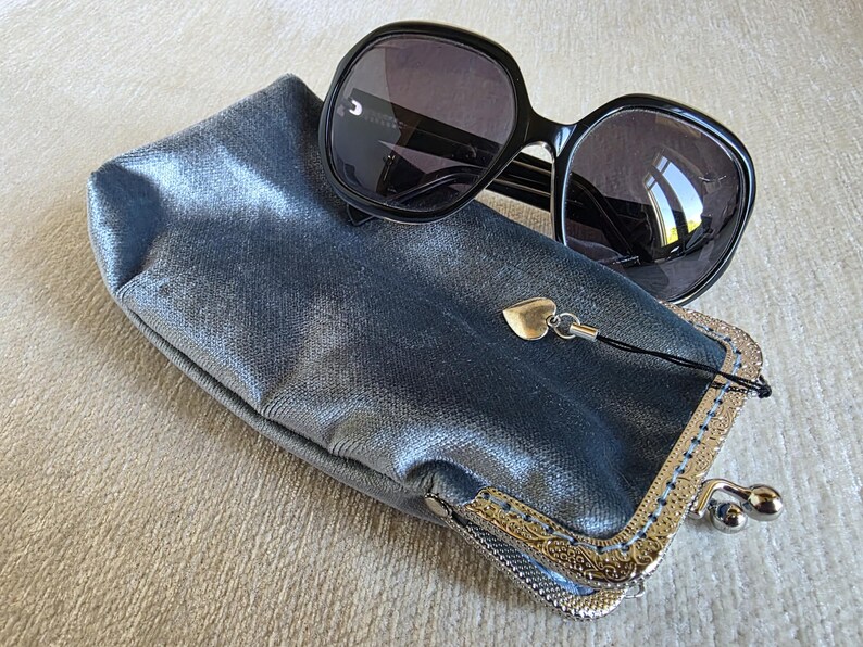 Puede incluir: Un par de gafas de sol negras de gran tama&ntilde;o sobre un estuche rectangular de color azul met&aacute;lico. El estuche cuenta con un cierre de boquilla plateado con un patr&oacute;n grabado ornamentado y un peque&ntilde;o dije de coraz&oacute;n plateado colgante.