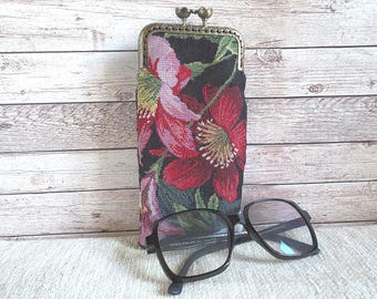 May include: A rectangular fabric spectacle case featuring a woven floral pattern in shades of red, pink, and green on a black background, finished with a bronze-toned metal kiss-lock clasp, positioned behind a pair of black-framed spectacles.