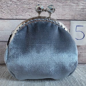 May include: A light blue velvet coin purse with a silver-toned metal kiss-lock frame. The clasp features two round, clear decorative knobs. A small white tag with the number 5 printed in blue is positioned to the right of the purse.