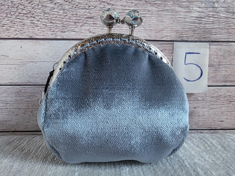 May include: A light blue velvet coin purse with a silver-toned metal kiss-lock frame. The clasp features two round, clear decorative knobs. A small white tag with the number 5 printed in blue is positioned to the right of the purse.