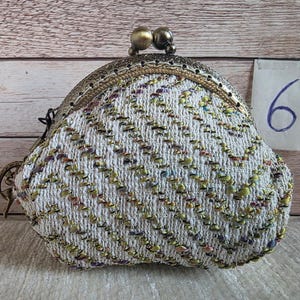 May include: A coin purse featuring a white textured fabric with a chevron pattern in green, purple, and gold metallic threads. The purse has an ornate antique brass-toned metal frame with a double ball clasp and a small tag labelled with the number 6.