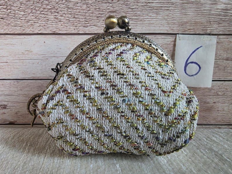 May include: A coin purse featuring a white textured fabric with a chevron pattern in green, purple, and gold metallic threads. The purse has an ornate antique brass-toned metal frame with a double ball clasp and a small tag labelled with the number 6.