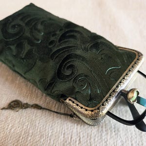 Puede incluir: Un estuche para gafas de tela verde oscuro con un patr&oacute;n de remolinos en relieve y un marco met&aacute;lico con un dise&ntilde;o de filigrana ornamentado. Un par de gafas con montura negra est&aacute; parcialmente insertado en el estuche, que incluye un peque&ntilde;o dije.