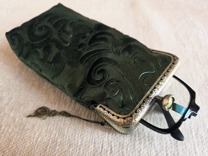 Puede incluir: Un estuche para gafas de tela verde oscuro con un patr&oacute;n de remolinos en relieve y un marco met&aacute;lico con un dise&ntilde;o de filigrana ornamentado. Un par de gafas con montura negra est&aacute; parcialmente insertado en el estuche, que incluye un peque&ntilde;o dije.