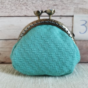 May include: A teal coin purse with a textured, woven fabric pattern and a bronze-toned metal frame. The clasp features two small, rounded floral-shaped knobs at the top. A small white tag with the handwritten number 3 is visible in the background.