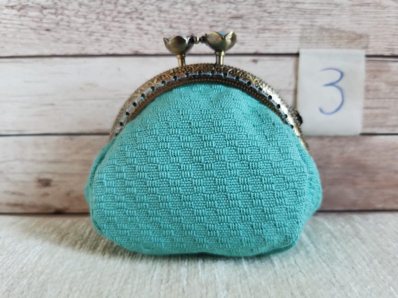 May include: A teal coin purse with a textured, woven fabric pattern and a bronze-toned metal frame. The clasp features two small, rounded floral-shaped knobs at the top. A small white tag with the handwritten number 3 is visible in the background.