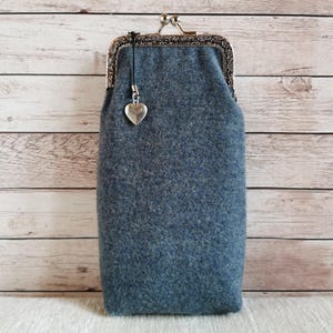 Puede incluir: Un estuche de lana afieltrada de color azul gris&aacute;ceo con forma rectangular y un cierre de boquilla de metal plateado. Un peque&ntilde;o dije plateado en forma de coraz&oacute;n cuelga de un cord&oacute;n negro unido al cierre superior del estuche de tela texturizada.