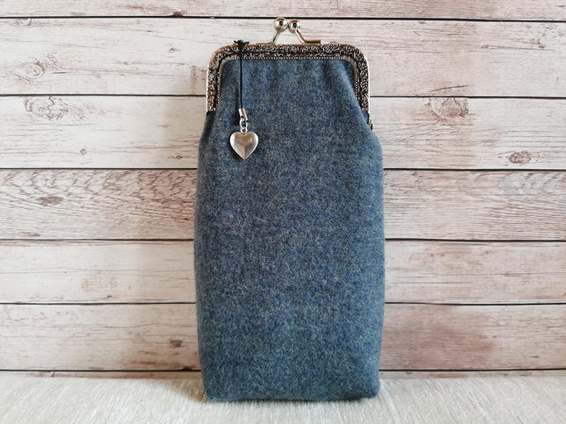 Puede incluir: Un estuche de lana afieltrada de color azul gris&aacute;ceo con forma rectangular y un cierre de boquilla de metal plateado. Un peque&ntilde;o dije plateado en forma de coraz&oacute;n cuelga de un cord&oacute;n negro unido al cierre superior del estuche de tela texturizada.