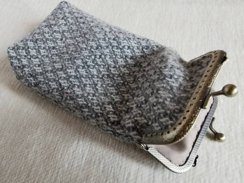 Puede incluir: Un estuche rectangular hecho de tela de tweed moteada en gris y blanco. El estuche cuenta con un marco de metal con cierre de boquilla en tono lat&oacute;n antiguo, con un patr&oacute;n decorativo en relieve y dos pomos esf&eacute;ricos en la parte superior.