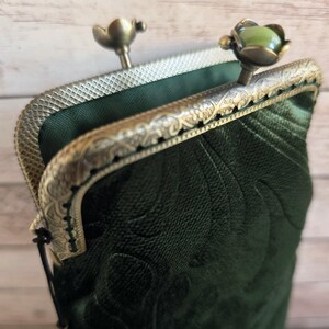 Puede incluir: Un bolso de terciopelo verde oscuro con un patr&oacute;n floral en relieve y un marco de metal en tono bronce. El marco presenta un dise&ntilde;o grabado intrincado y dos cierres, uno de los cuales est&aacute; rematado con una piedra redonda, lisa y verde claro.