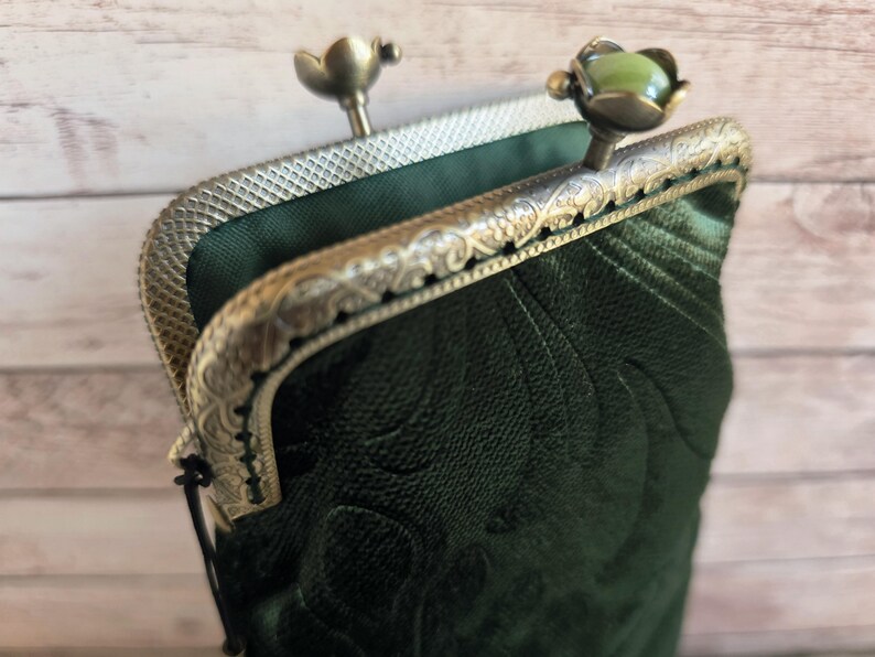 Puede incluir: Un bolso de terciopelo verde oscuro con un patr&oacute;n floral en relieve y un marco de metal en tono bronce. El marco presenta un dise&ntilde;o grabado intrincado y dos cierres, uno de los cuales est&aacute; rematado con una piedra redonda, lisa y verde claro.