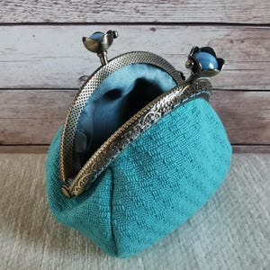 May include: A teal coin purse with a textured fabric exterior and a smooth teal interior lining. The purse features an antique bronze-toned metal frame with an ornate embossed pattern and two clasp knobs topped with smooth, round, light blue beads.
