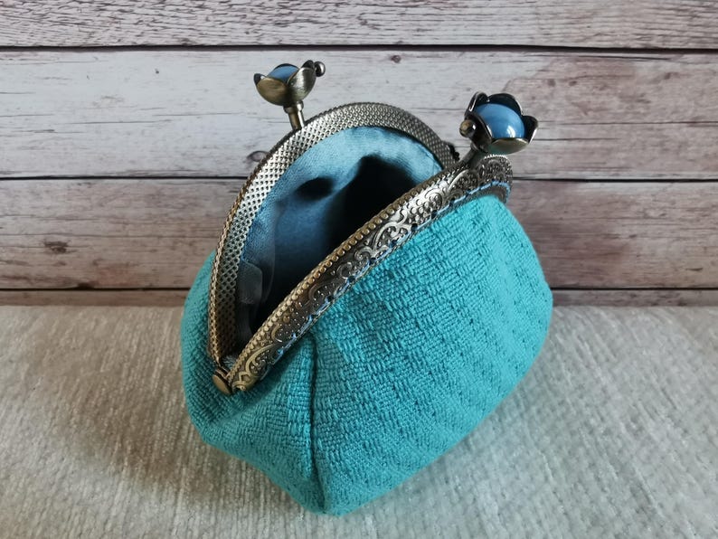 May include: A teal coin purse with a textured fabric exterior and a smooth teal interior lining. The purse features an antique bronze-toned metal frame with an ornate embossed pattern and two clasp knobs topped with smooth, round, light blue beads.