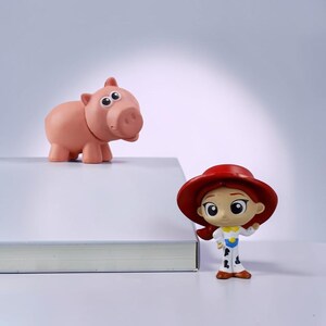 May include: A small pink pig figurine with large eyes and a rounded snout positioned next to a small figurine of a person wearing a wide-brimmed red hat, a yellow shirt, and white pants with black cow-print spots, both standing near a white book.