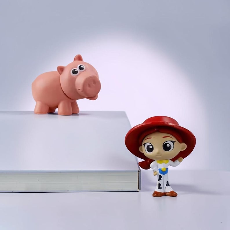 May include: A small pink pig figurine with large eyes and a rounded snout positioned next to a small figurine of a person wearing a wide-brimmed red hat, a yellow shirt, and white pants with black cow-print spots, both standing near a white book.