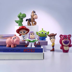 May include: A collection of small plastic character figurines including a green dinosaur, a brown horse, a red-hatted cowgirl, a space ranger, a pink pig, a three-eyed green alien, a cowboy, and a purple bear, arranged on stacked books.