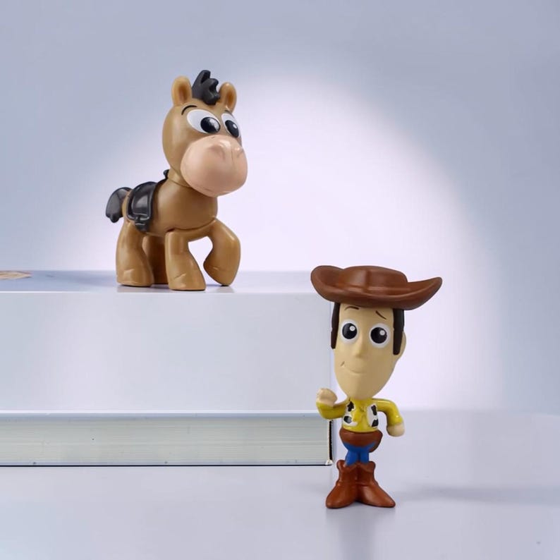 May include: Two plastic figurines featuring a tan horse with a black saddle and a character wearing a brown cowboy hat, yellow shirt with cow print vest, blue pants, and brown boots. The horse stands on a white surface, while the character stands.