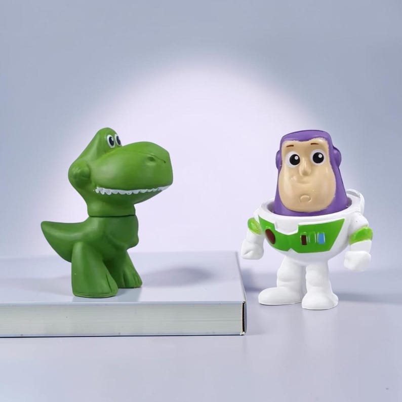 May include: Two small plastic character figurines. One is a green dinosaur with a large head and visible teeth. The other is a space ranger character with a purple hood, a white suit featuring green accents, and a round chest plate with small buttons.