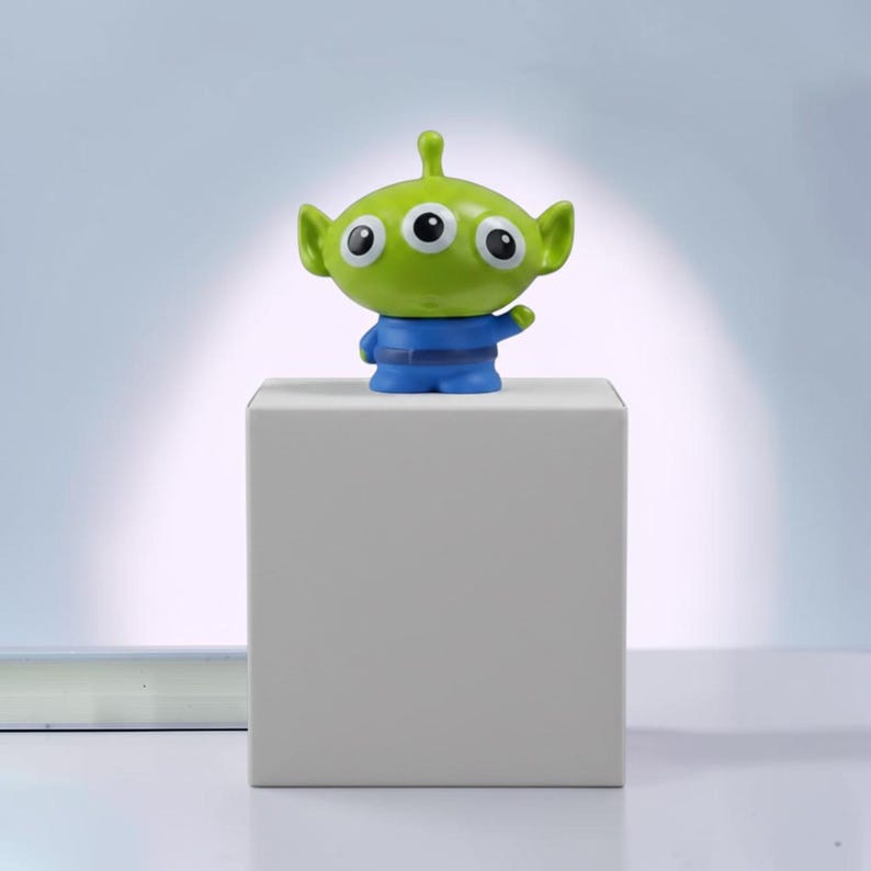 May include: A small green alien figurine with three eyes, a pointed antenna on its head, and a blue uniform. The character has a wide head, large black pupils, and a small mouth. It is positioned standing upright on a plain white square block.