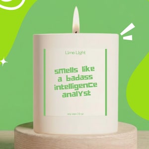 May include: A white cylindrical soy wax candle with a lit wick. The label features the text Lime Light, smells like a badass intelligence analyst, and soy wax | 9 oz in green font, framed by two vertical green lines on a white rectangular background.