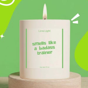 May include: A white cylindrical candle with a lit wick, featuring a label that reads Lime Light, smells like a badass trainer, and soy wax 9 oz. The candle is positioned on a light-colored wooden base against a bright green background.