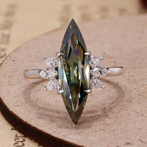 May include: A silver-toned ring featuring a large, elongated marquise-cut dark green gemstone as the centerpiece. The central stone is flanked on both sides by three small, clear, pear-shaped accent stones set into the polished metal band.