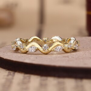 May include: A gold band ring featuring a continuous wavy design. The ring is set with alternating marquise-cut and round-cut clear stones embedded along the undulating surface of the metal band, creating a repeating pattern of sparkling accents.