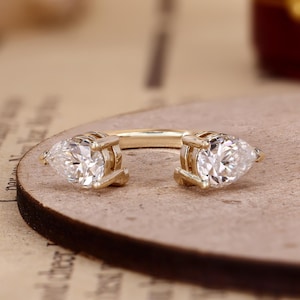 May include: A gold open-style ring featuring two pear-cut clear stones, one at each end of the band. The stones are held in place by four-prong settings, creating a symmetrical design with a polished metallic finish and clear faceted gems.