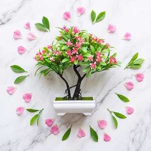 Pode incluir: Um pequeno bonsai artificial com ramos castanho-escuros, folhas verde-vibrantes e cachos de pequenas flores cor-de-rosa. A árvore está plantada num recipiente retangular de cerâmica branca, rodeada por pétalas cor-de-rosa e folhas verdes.