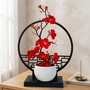 Può includere: Un vaso in ceramica bianca contenente un ramo con fiori rosso vivo, posizionato davanti a una cornice circolare in metallo nero con base a griglia geometrica, montato su una solida base rettangolare in legno nero.