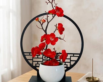 Peut inclure: Un pot en céramique blanche contenant une branche aux fleurs rouge vif, placé devant un cadre métallique circulaire noir avec une base en treillis géométrique, le tout monté sur un socle rectangulaire en bois noir massif.