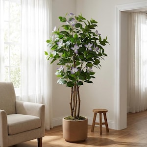Könnte beinhalten: Ein hoher künstlicher Baum mit mehreren dünnen braunen Stämmen und üppigem grünen Blattwerk. Der Baum weist mehrere hellviolette, vierblättrige Blüten auf, die über das Laub verteilt sind. Er steht in einem schlichten, zylindrischen beigen Topf.