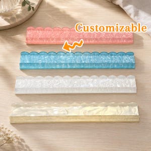 Può includere: Quattro ripiani espositivi rettangolari in acrilico con bordi superiori smerlati, disposti verticalmente. I ripiani presentano finiture marmorizzate rosa, blu, bianco e crema. Il testo "Customizable" appare in arancione sopra il ripiano blu.