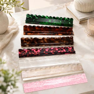May include: A set of seven rectangular hair clips arranged in a vertical stack. The clips feature a scalloped top edge and come in various colours and patterns, including green, brown, and black tortoiseshell, pink, clear, and cream marble finishes.