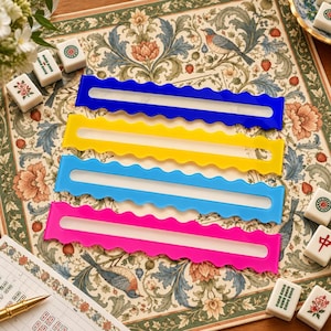 Pode incluir: Quatro suportes retangulares para peças de mahjong com bordas onduladas, nas cores azul royal, amarelo, azul claro e rosa choque. Cada suporte possui uma longa fenda horizontal para segurar as peças, dispostos verticalmente sobre uma superfície com estampa floral.