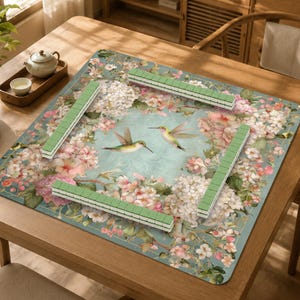 May include: A square mahjong mat featuring a light blue background with a floral pattern of white and pink blossoms and two hummingbirds in the center. Four rows of green mahjong tiles are arranged in a square formation on the mat surface.
