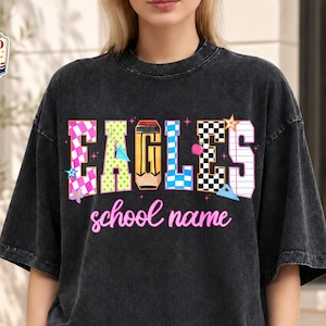 May include: A black mineral-washed t-shirt featuring the word EAGLES in colorful, patterned block letters, including a pencil design for the letter L. Below the word, the text school name is printed in a cursive pink font with small stars.