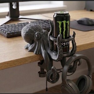 May include: A dark grey octopus-shaped desk accessory featuring a circular holder for a beverage can and a hook for hanging headphones. The octopus is secured to the edge of a desk with a screw-clamp mechanism, with tentacles extending to support the items.