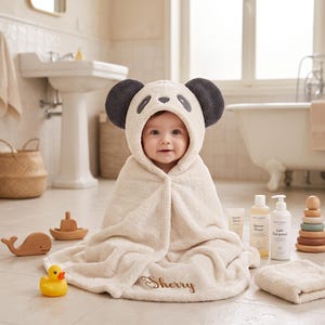 Puede incluir: Una toalla de baño con capucha de color crema que presenta un diseño de cara de panda con orejas gris oscuro y detalles bordados. La toalla está personalizada con el nombre Sherry en letra cursiva en el borde inferior, de textura suave.
