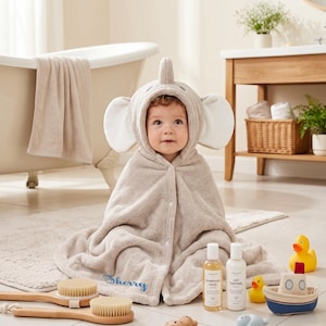 Peut inclure: Une serviette de bain à capuche pour bébé de couleur beige, ornée d'un motif d'éléphant avec de grandes oreilles et une trompe sur la capuche. Le tissu doux et texturé présente une fermeture à pression et le prénom Sherry brodé en bleu.